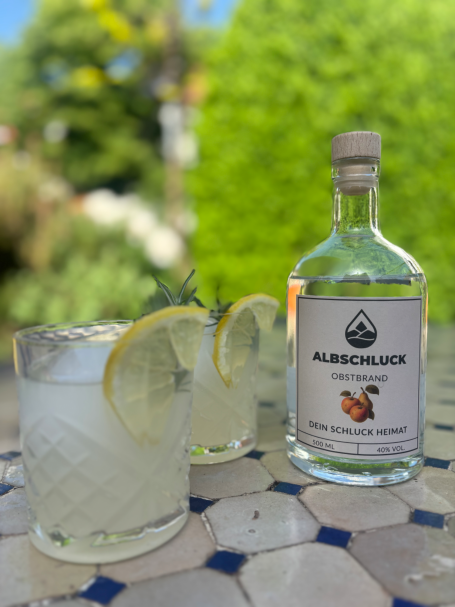 Zwei Tumblergläser dekoriert mit Zitrone und Rosmarin, daneben eine Flasche klarer Albschluck Obstbrand auf einem Tisch im Garten.